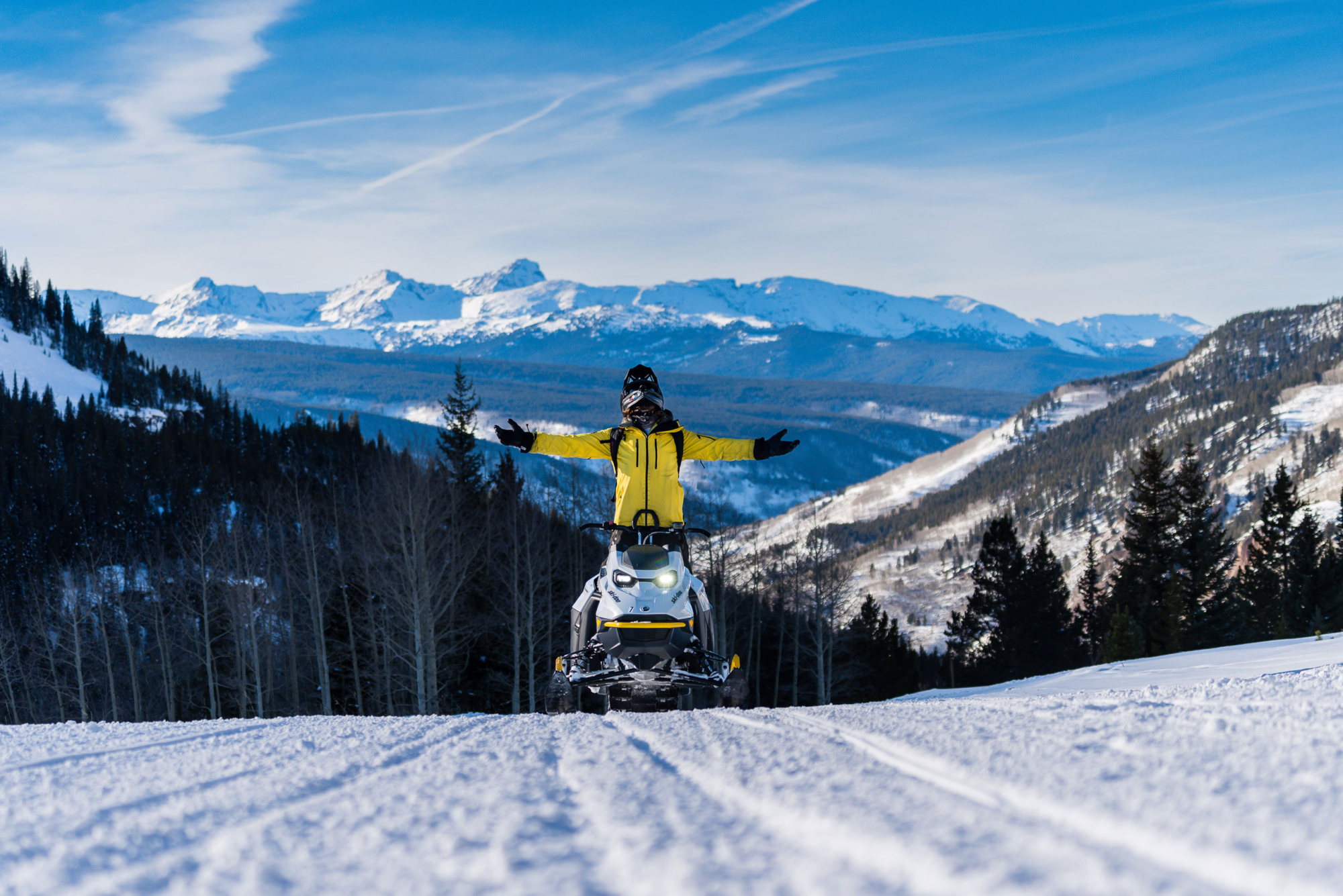 A person standing on a snowmobile at the top of a snow covered slope.